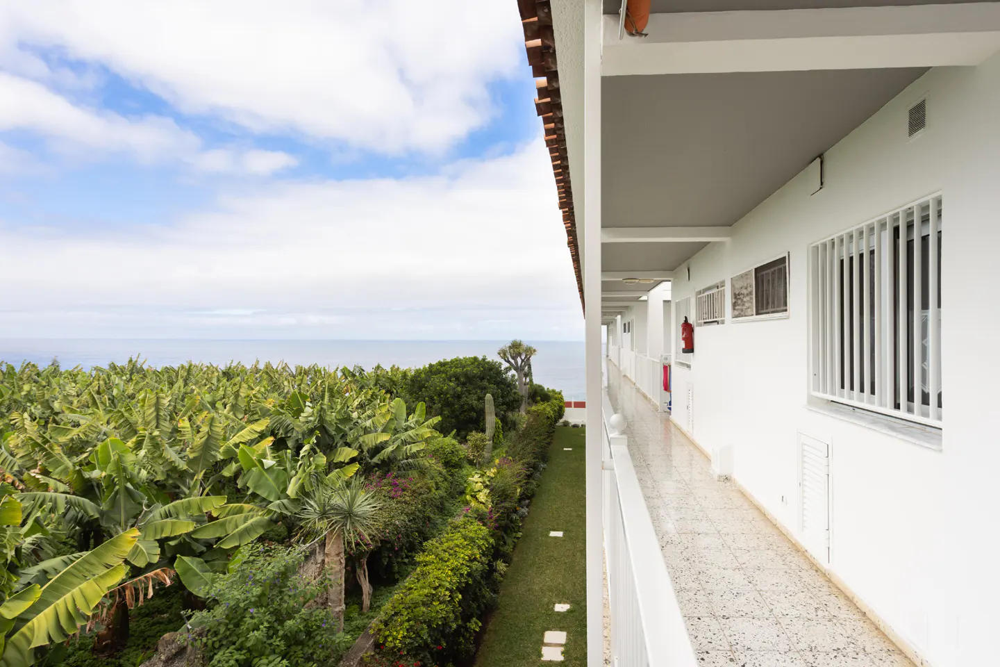 Balcony view of a white building overlooking a lush green landscape and the ocean under a cloudy sky.