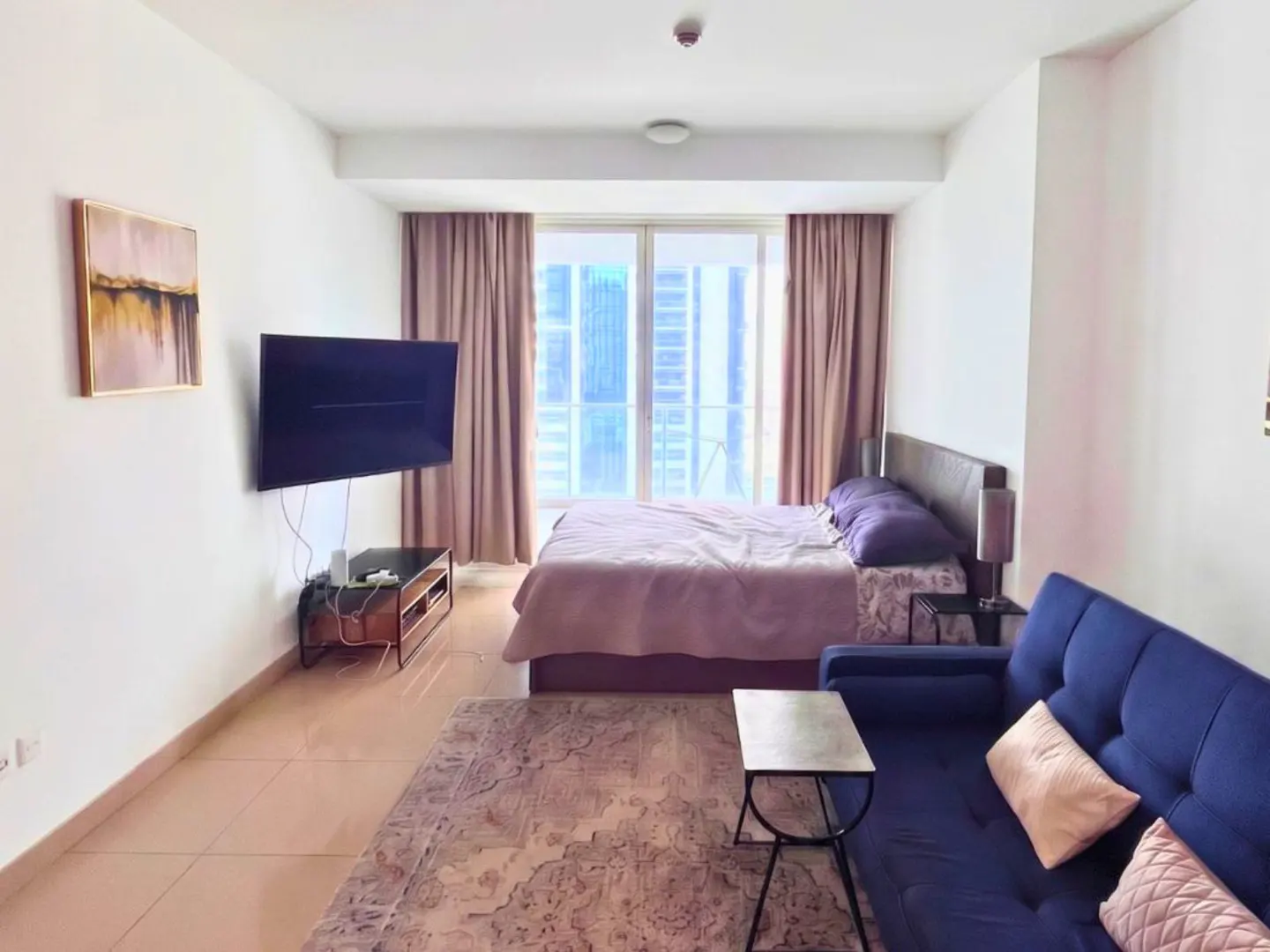 Studio apartment with a blue sofa, bed, TV, and a balcony with city views. The walls are white, and the floor is tiled.