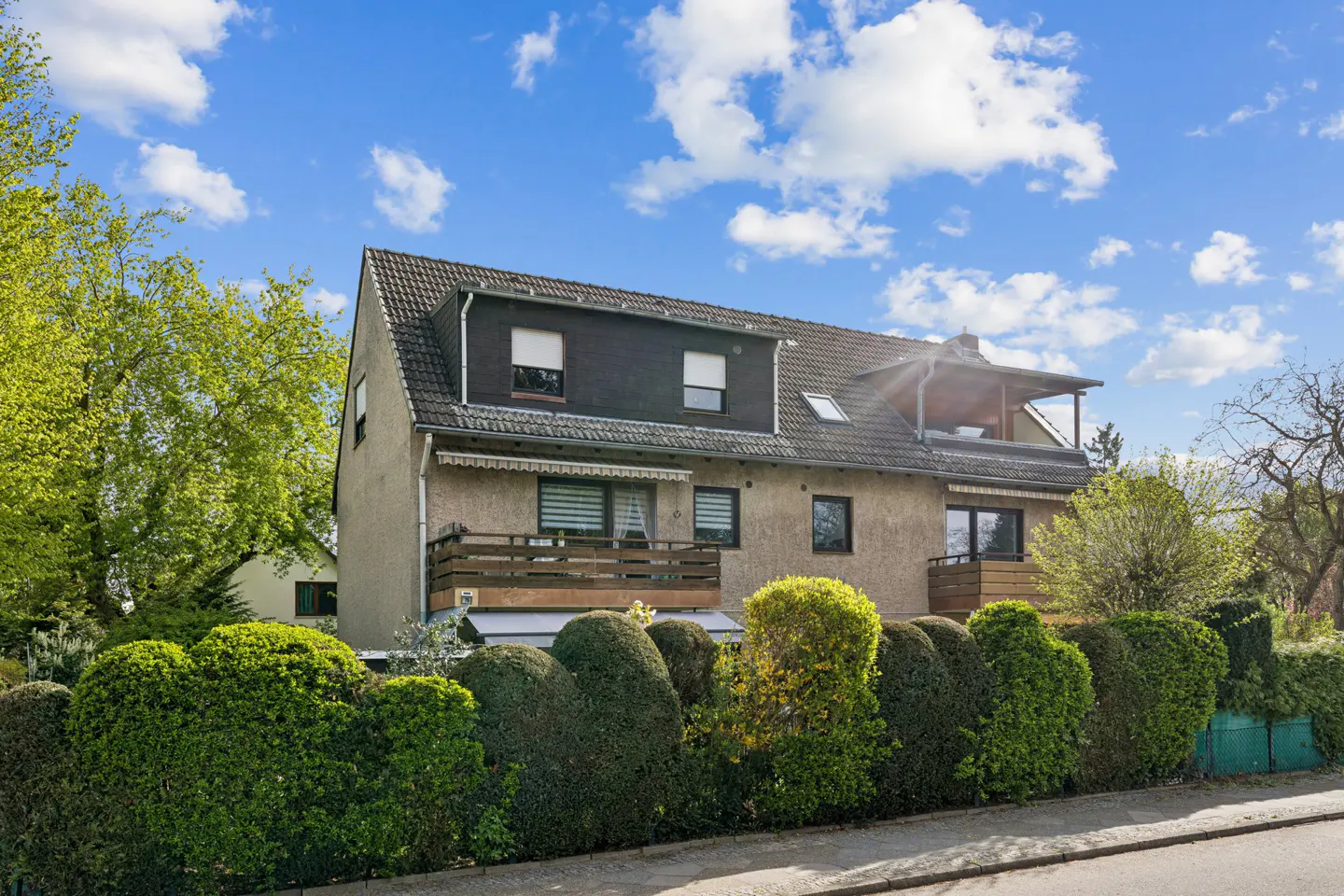 Two-story beige house with a dark roof, wooden balconies, and a manicured green hedge in front under a blue sky.