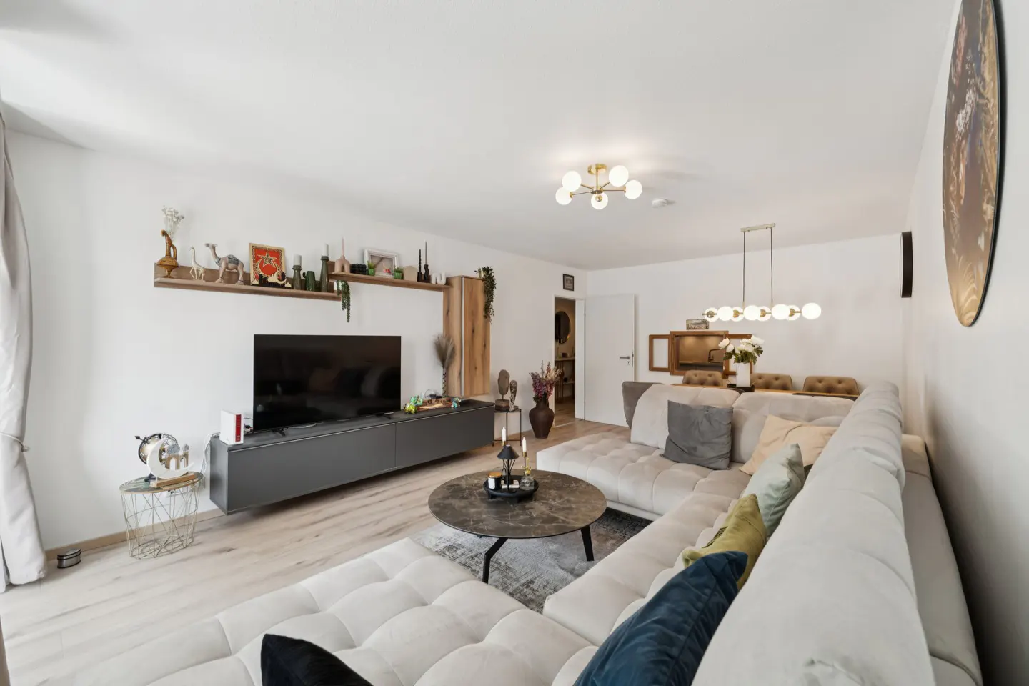 Bright living room with a large white sectional sofa, a gray TV stand, and a round coffee table on a gray rug. A dining area is visible in the background.
