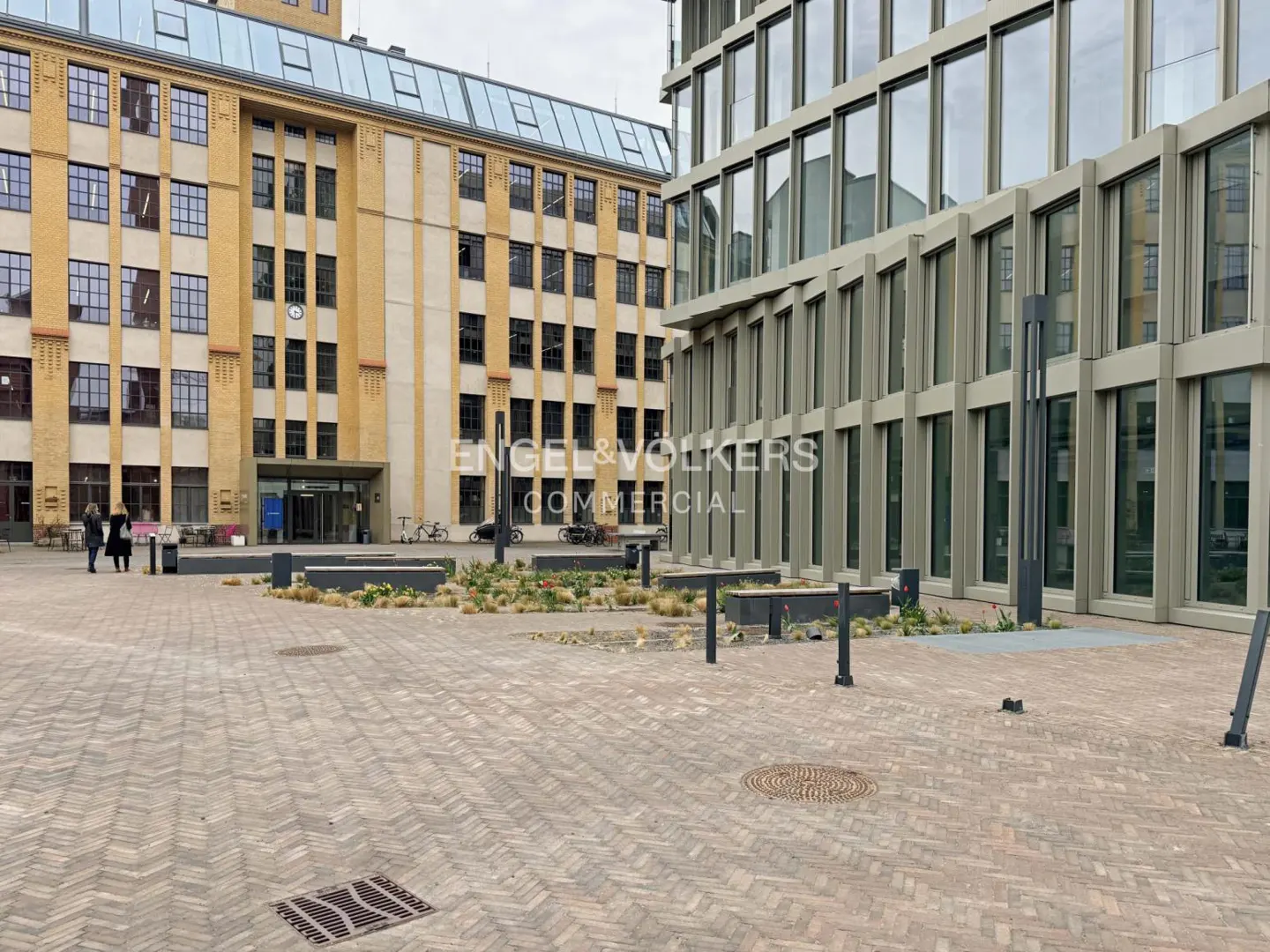 Exterior view of a modern office building with large windows and a brick courtyard. A yellow brick building is visible in the background.