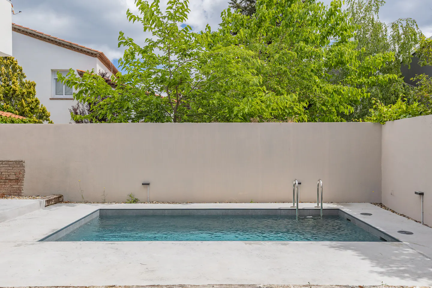 A backyard swimming pool with metal ladder, surrounded by a concrete patio and a tall beige wall. Green trees are visible above the wall.