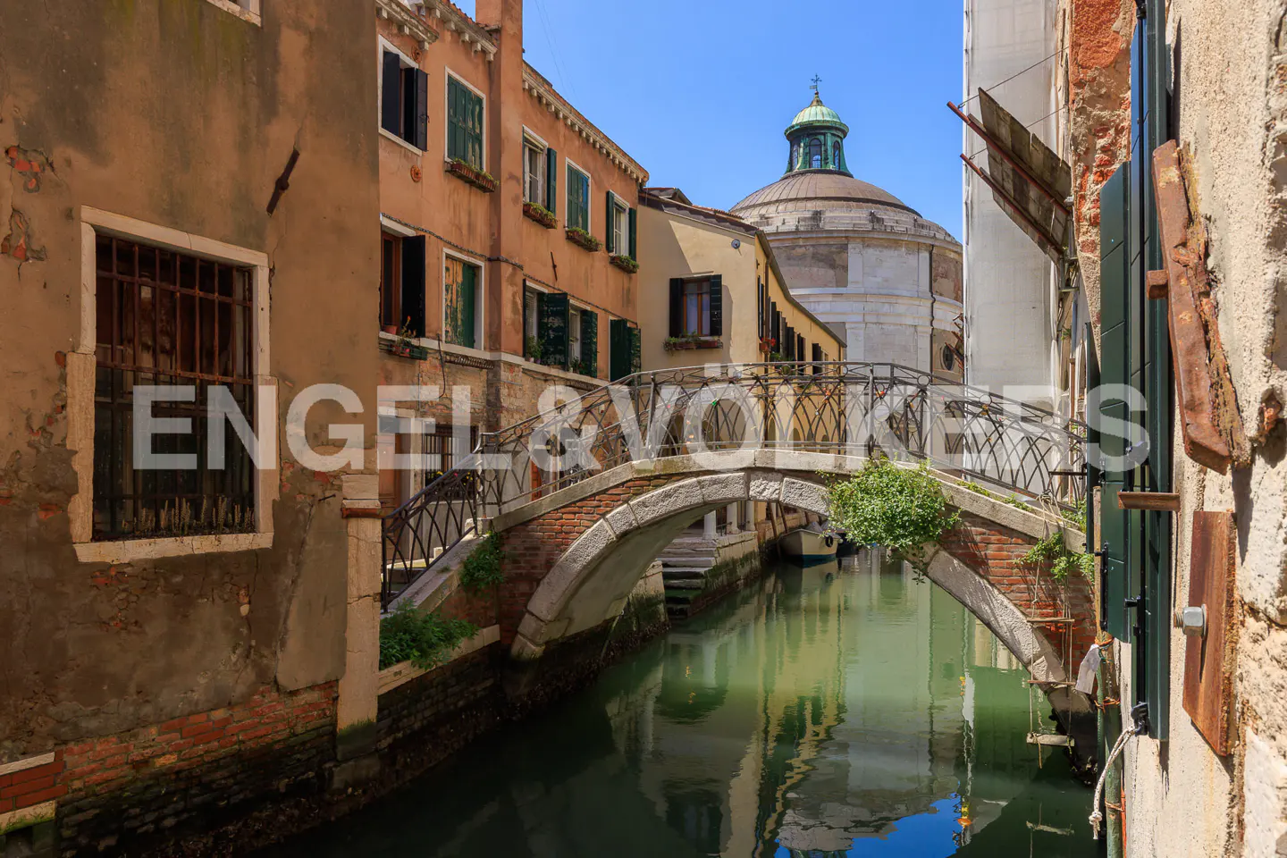 Venice canal view with a stone bridge, green water, and buildings with windows and shutters under a blue sky.