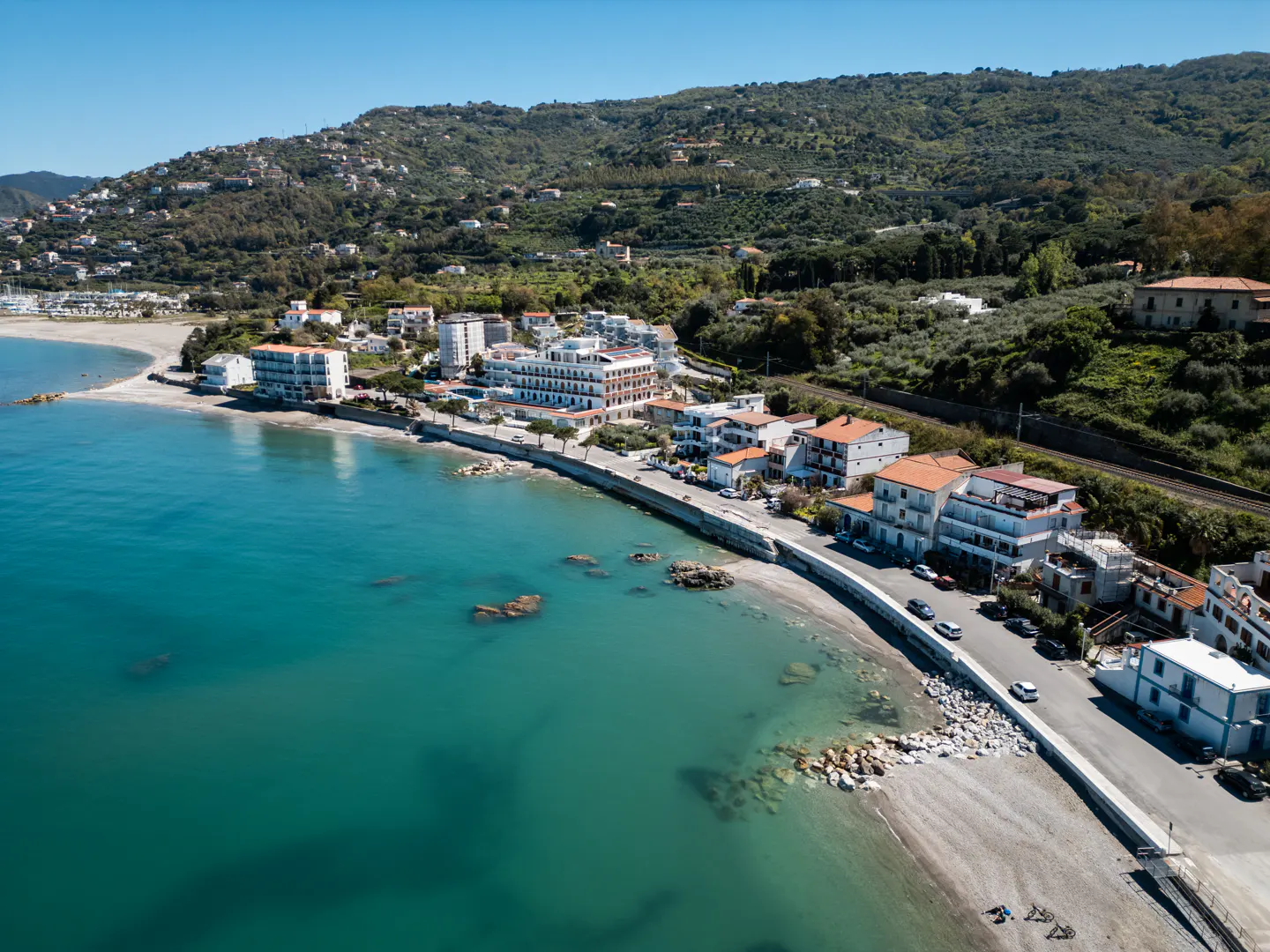 Aerial view of a coastal town with turquoise water, sandy beaches, buildings, and a green hillside in the background.