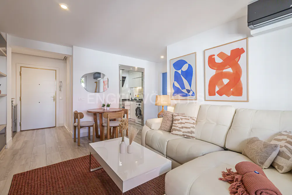 Bright living room with white walls, a white sofa, and abstract art. A round wooden table and chairs sit near a mirror. A white coffee table is in front of the sofa.