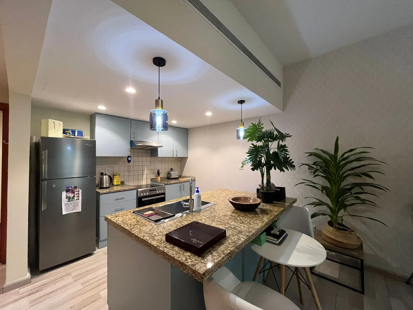Modern kitchen with light blue cabinets, granite island countertop, stainless steel fridge, and two pendant lights.
