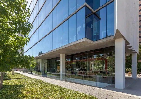 Modern office building with glass facade and concrete supports, surrounded by green landscaping and a paved walkway.