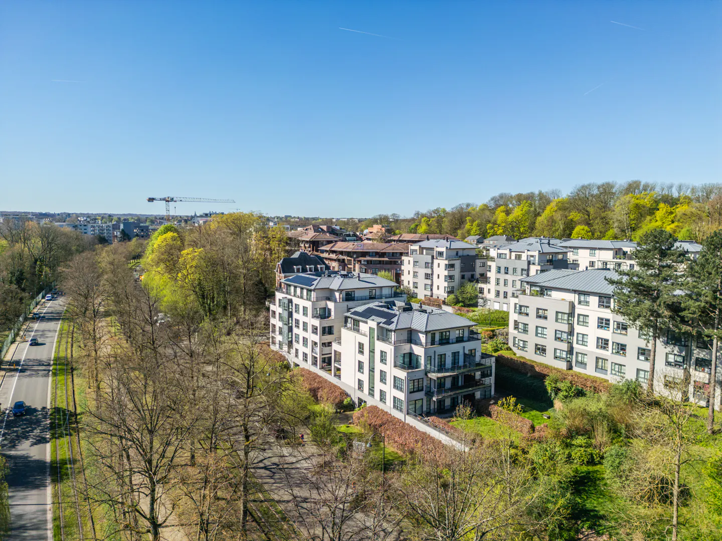 Aerial view of modern white apartment buildings with gray roofs, surrounded by green trees and a blue sky.
