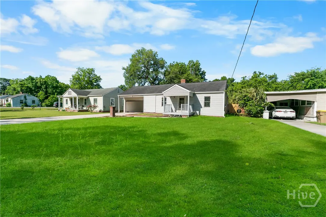 A light gray single-story house with a green lawn and a carport on a sunny day.