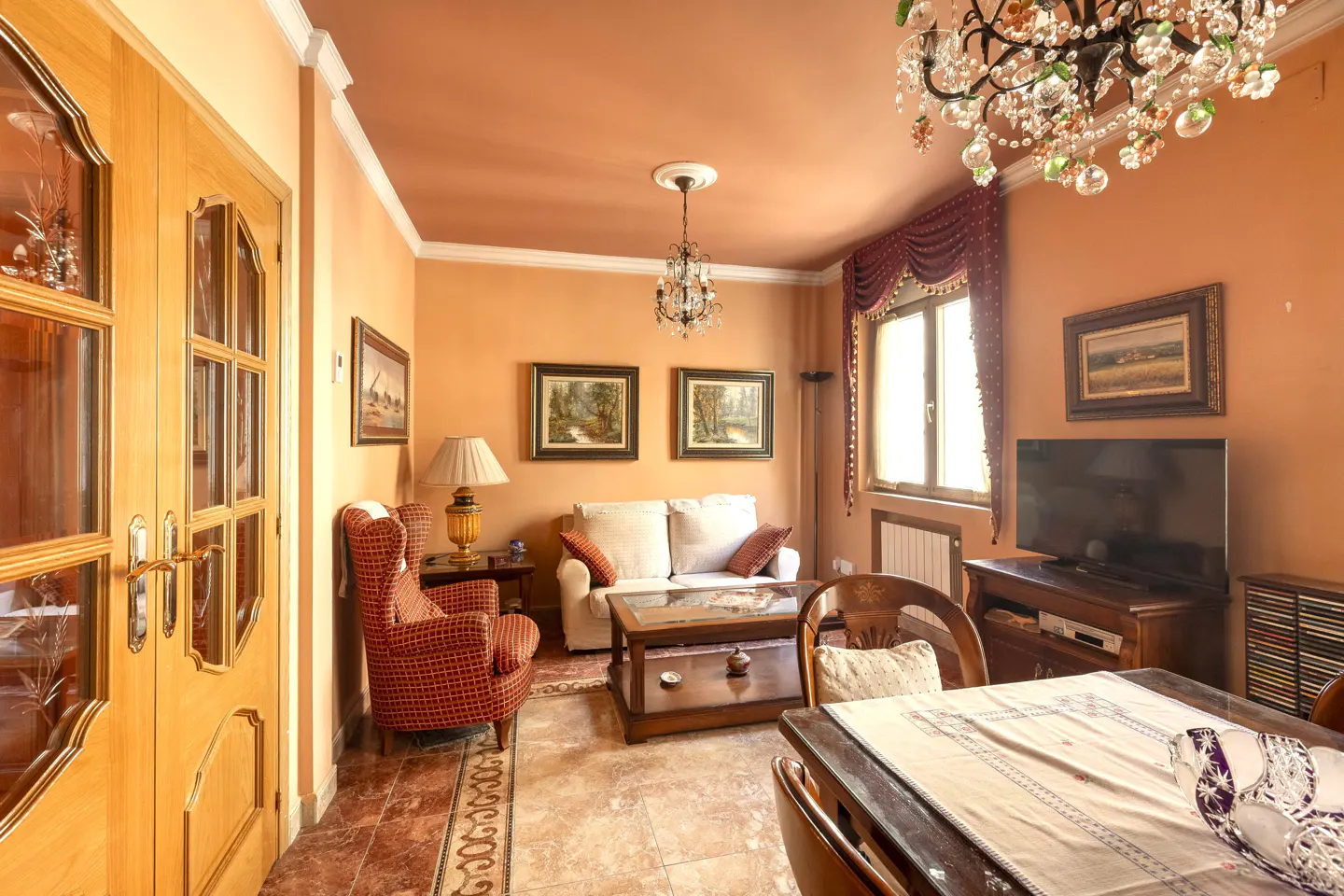 Living room with peach walls, tile floor, and dark wood furniture. A crystal chandelier hangs from the ceiling.