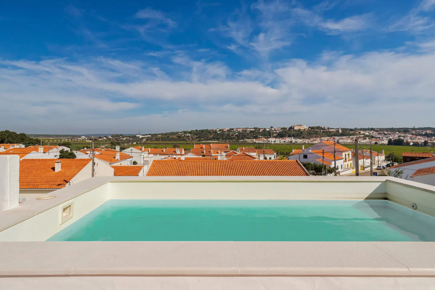 Rooftop pool with turquoise water overlooks a town with orange tile roofs under a blue, cloudy sky.