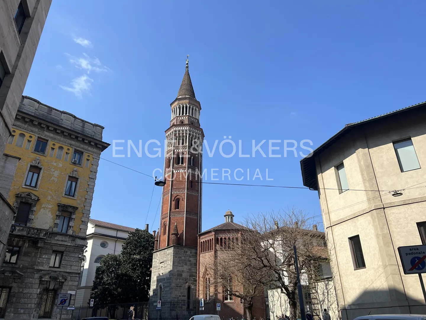 View of the Basilica di Sant'Eustorgio, a red brick church with a tall tower, surrounded by buildings under a blue sky.