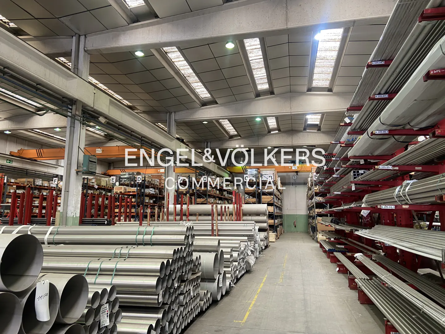 A warehouse interior with metal pipes stacked on shelves and on the floor. Overhead cranes are visible.