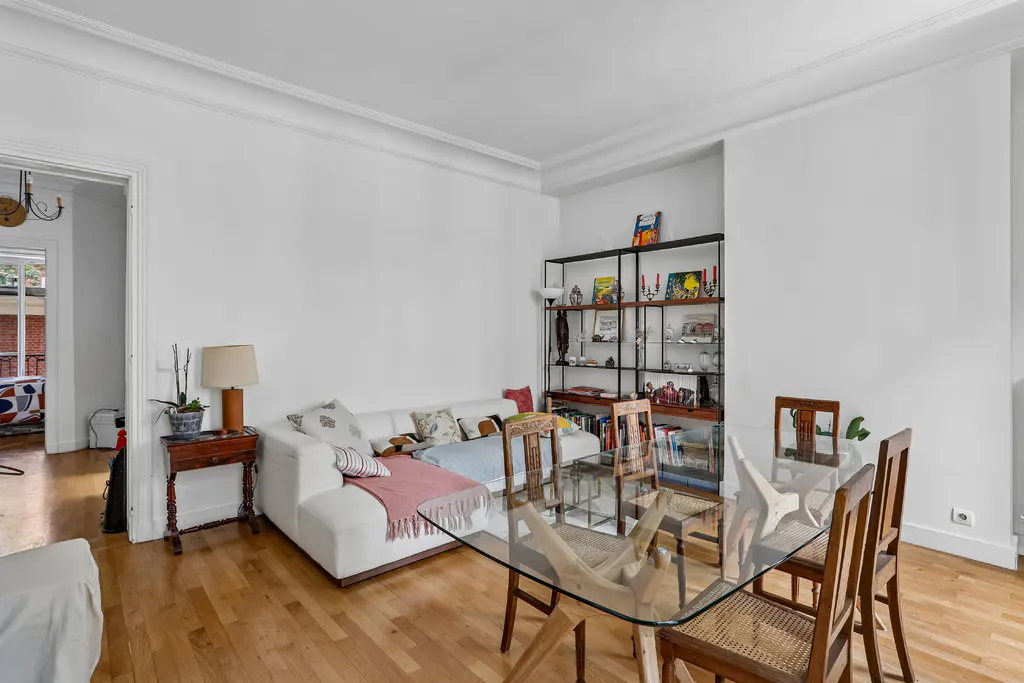 Bright living room with a white sofa, glass dining table, wooden chairs, and a tall metal shelving unit on hardwood floors.