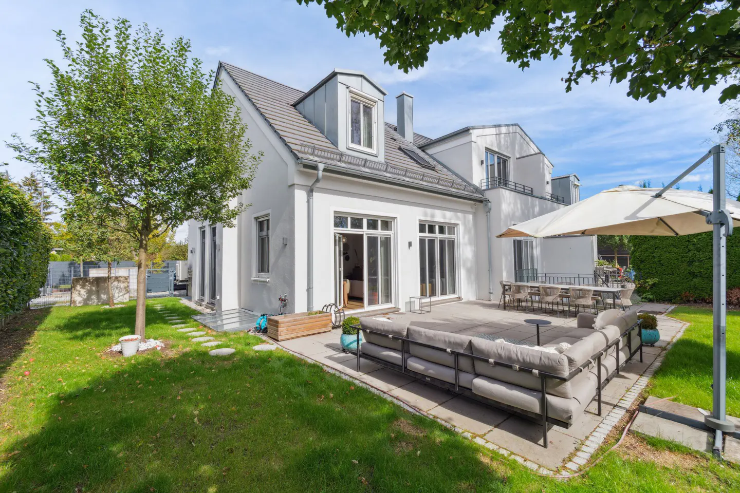 Backyard view of a white house with a gray patio, sofa, dining table, and a large umbrella.