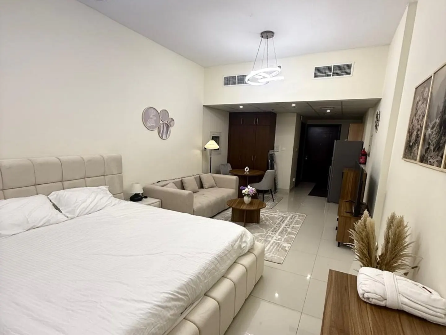 A bright studio apartment with a bed, sofa, table, and kitchen area. The walls are off-white, and the decor is modern.