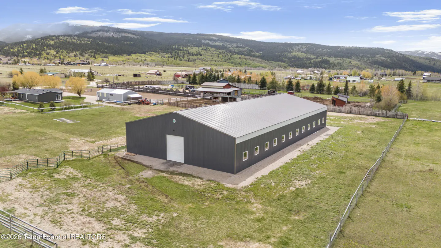 Aerial view of a large, dark grey metal barn in a grassy field with a mountain range in the background.
