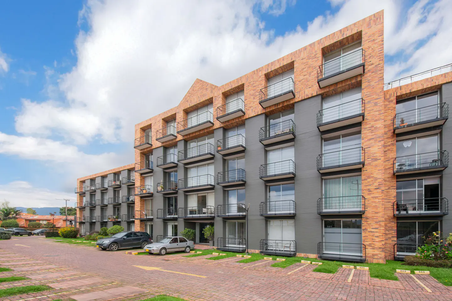 Exterior view of a modern gray and brick apartment building with black balconies and a brick parking lot.