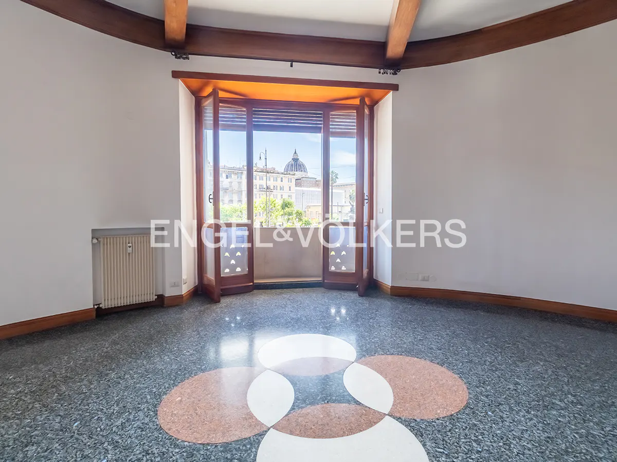 Empty room with white walls, wooden beams, and a balcony with a city view. The floor is terrazzo with a circular pattern.