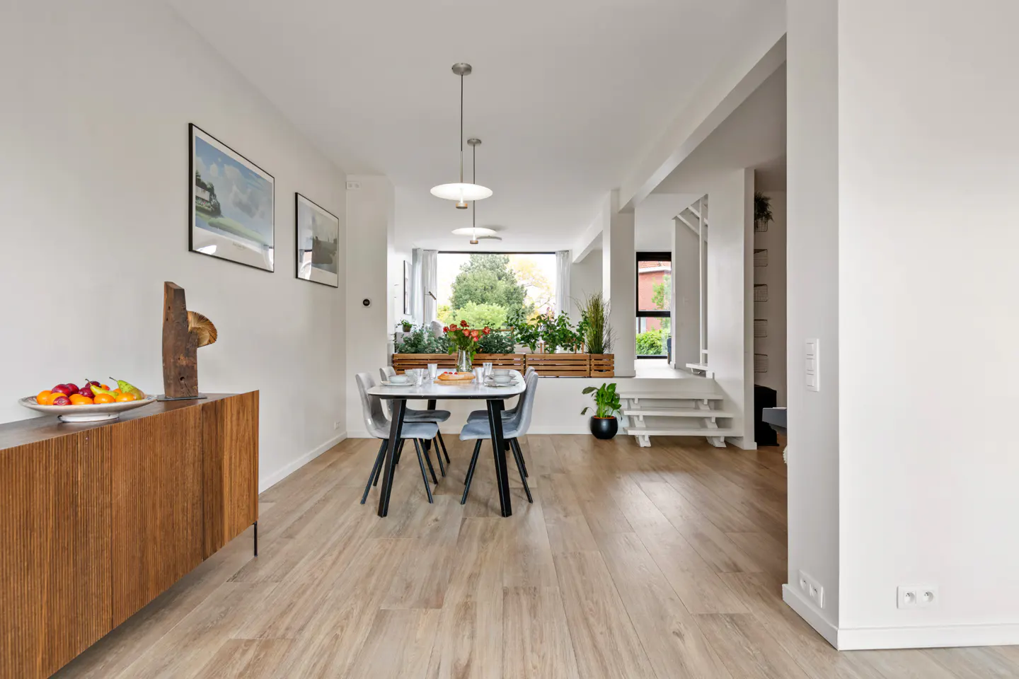 Bright dining room with wood floors, white walls, and a table with four chairs. A wooden cabinet holds fruit and art. Plants are visible through a large window.