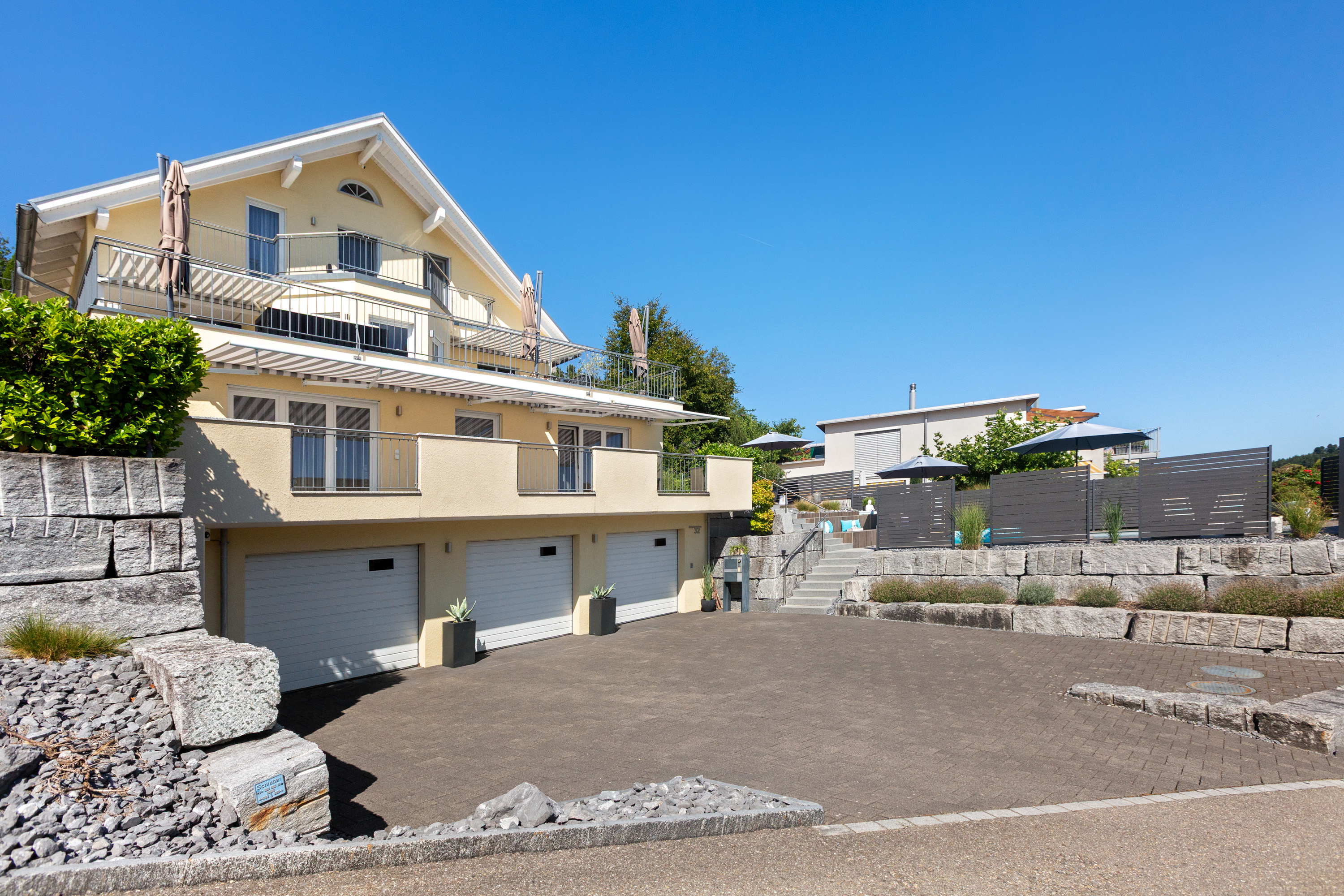 A light yellow three-story house with a triple garage, balconies, and a paved driveway under a clear blue sky. [AI-generated text]