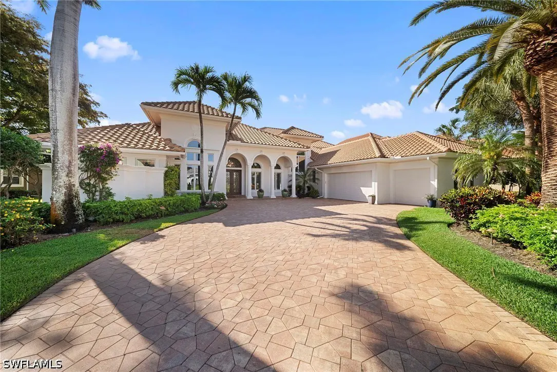 Mediterranean-style home with a tile roof, white exterior, arched entryway, and a patterned brick driveway. Palm trees and blue sky.