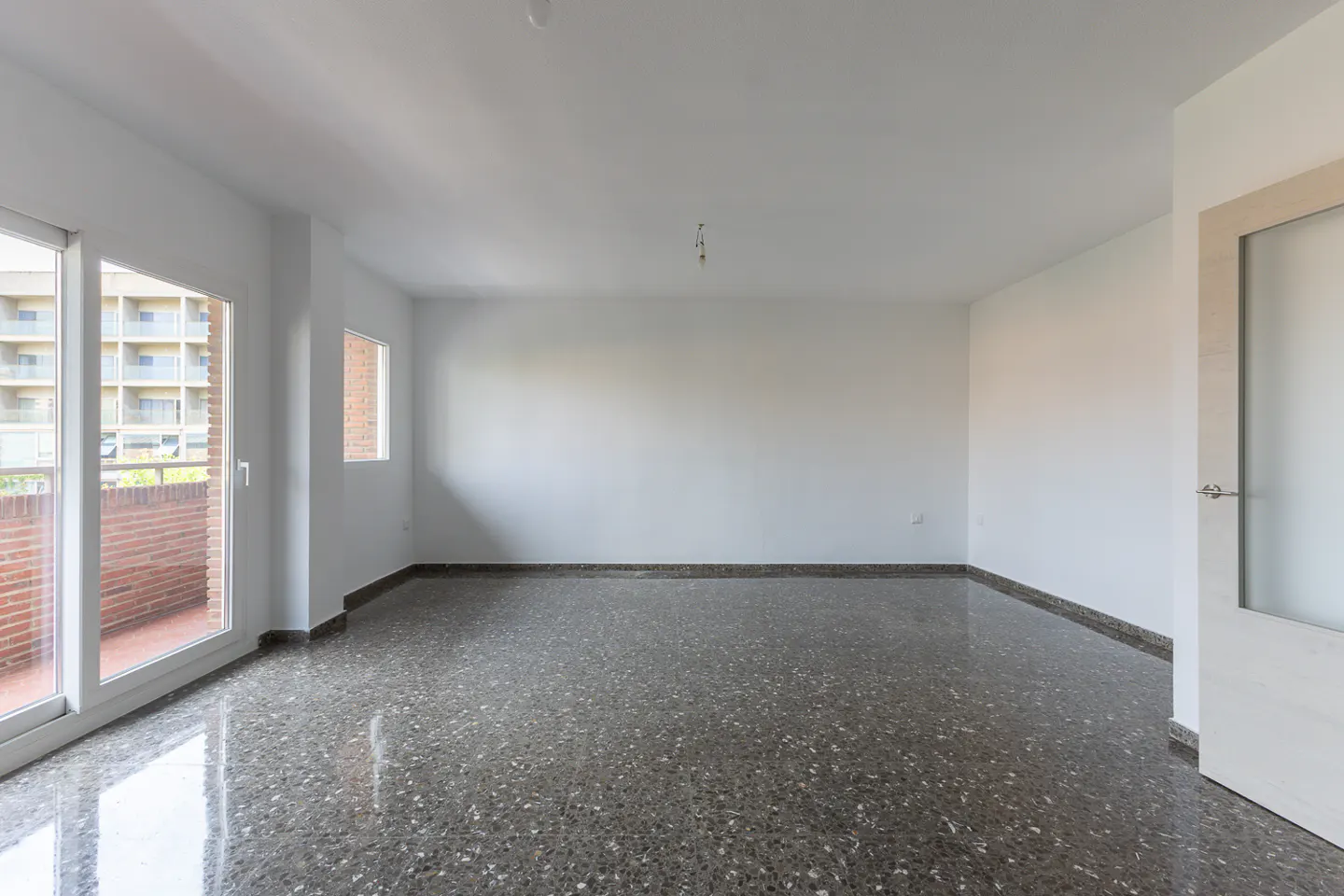 Empty room with white walls, polished dark grey terrazzo floors, and a glass door leading to a brick-walled balcony.
