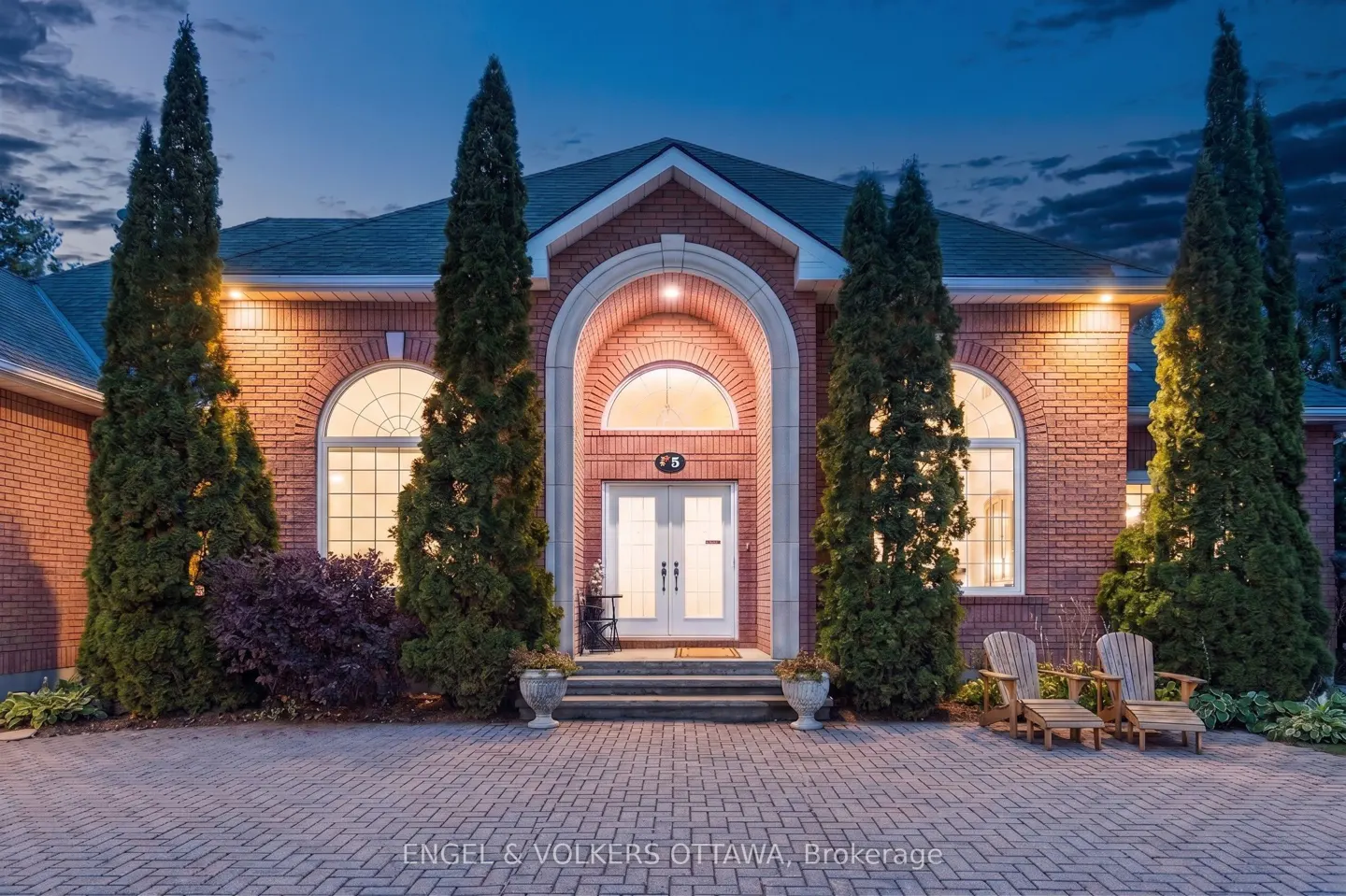 Brick house at dusk with arched entryway, white double doors, and tall evergreens. Two wooden chairs sit on the brick driveway.