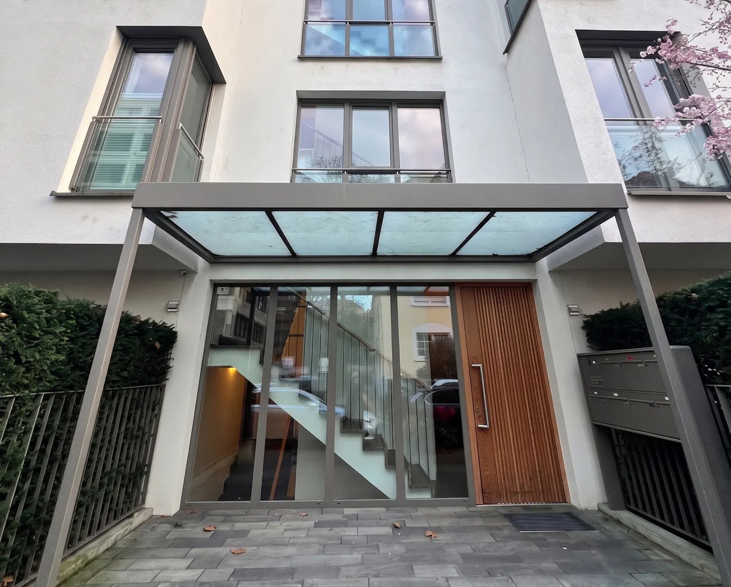 Modern apartment building entrance with a glass canopy, wooden door, and large windows revealing an interior staircase.