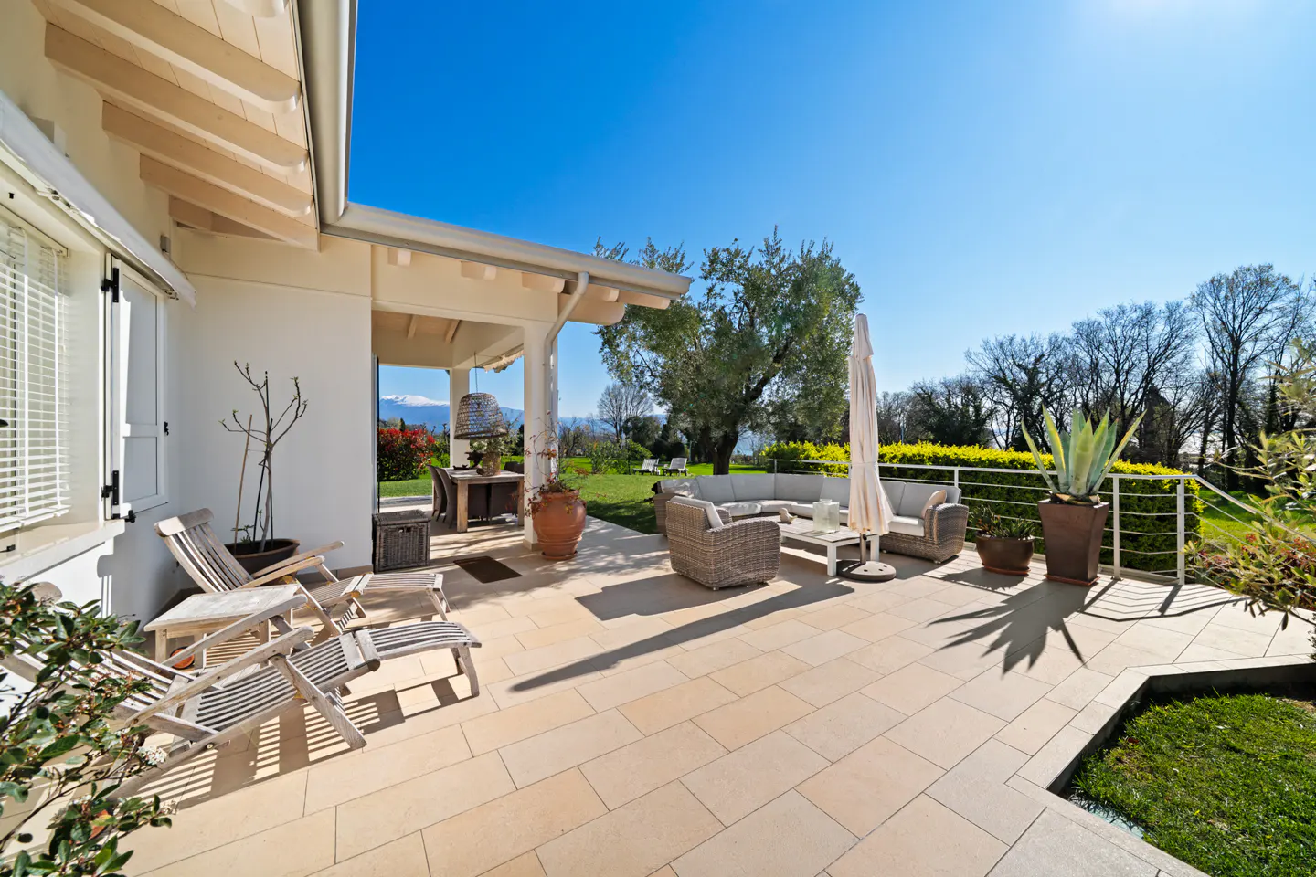 Outdoor patio with wicker furniture, lounge chairs, and a white umbrella. Green lawn and trees in the background under a clear blue sky.