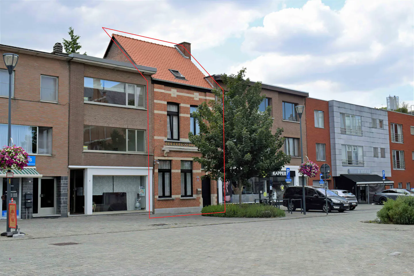 A red-brick townhouse with a tiled roof, outlined in red, situated in a row of buildings on a paved town square.