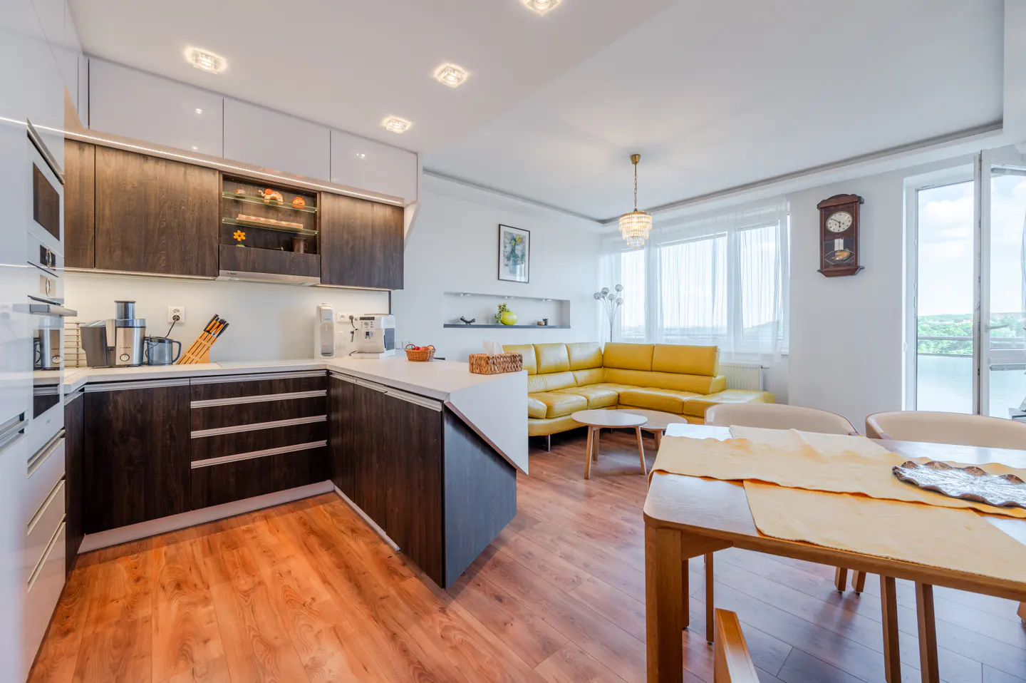 Bright, modern apartment interior with a dark wood kitchen, yellow sofa, and wooden dining table.