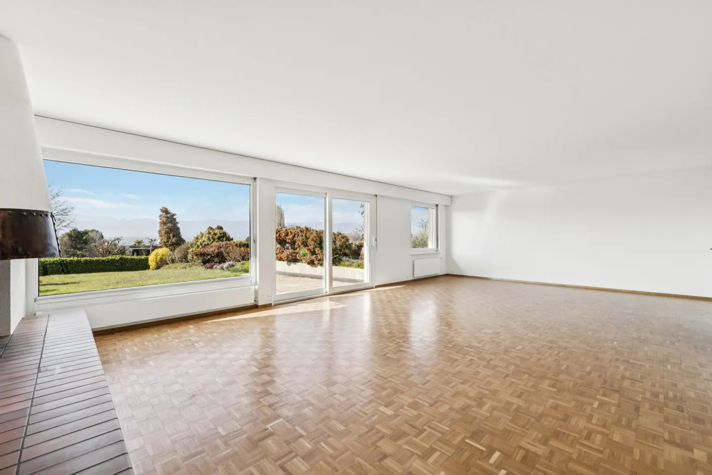 Bright, empty room with parquet floors, white walls, and large windows overlooking a green lawn and garden. A fireplace is visible on the left.