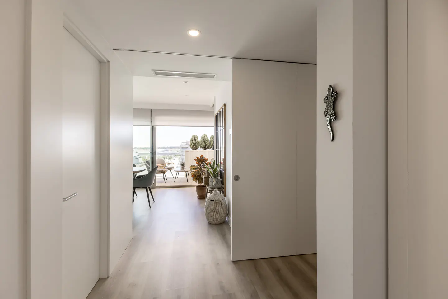A bright hallway with light wood floors leads to a sunlit living area and balcony with outdoor furniture and plants.