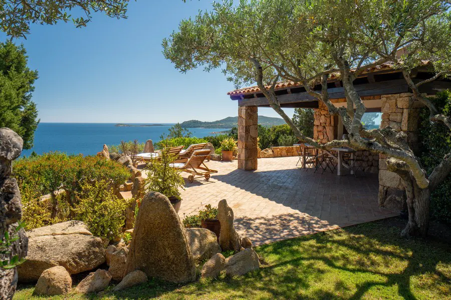 Outdoor patio with stone columns, lounge chairs, and ocean view. Green trees and blue sky.