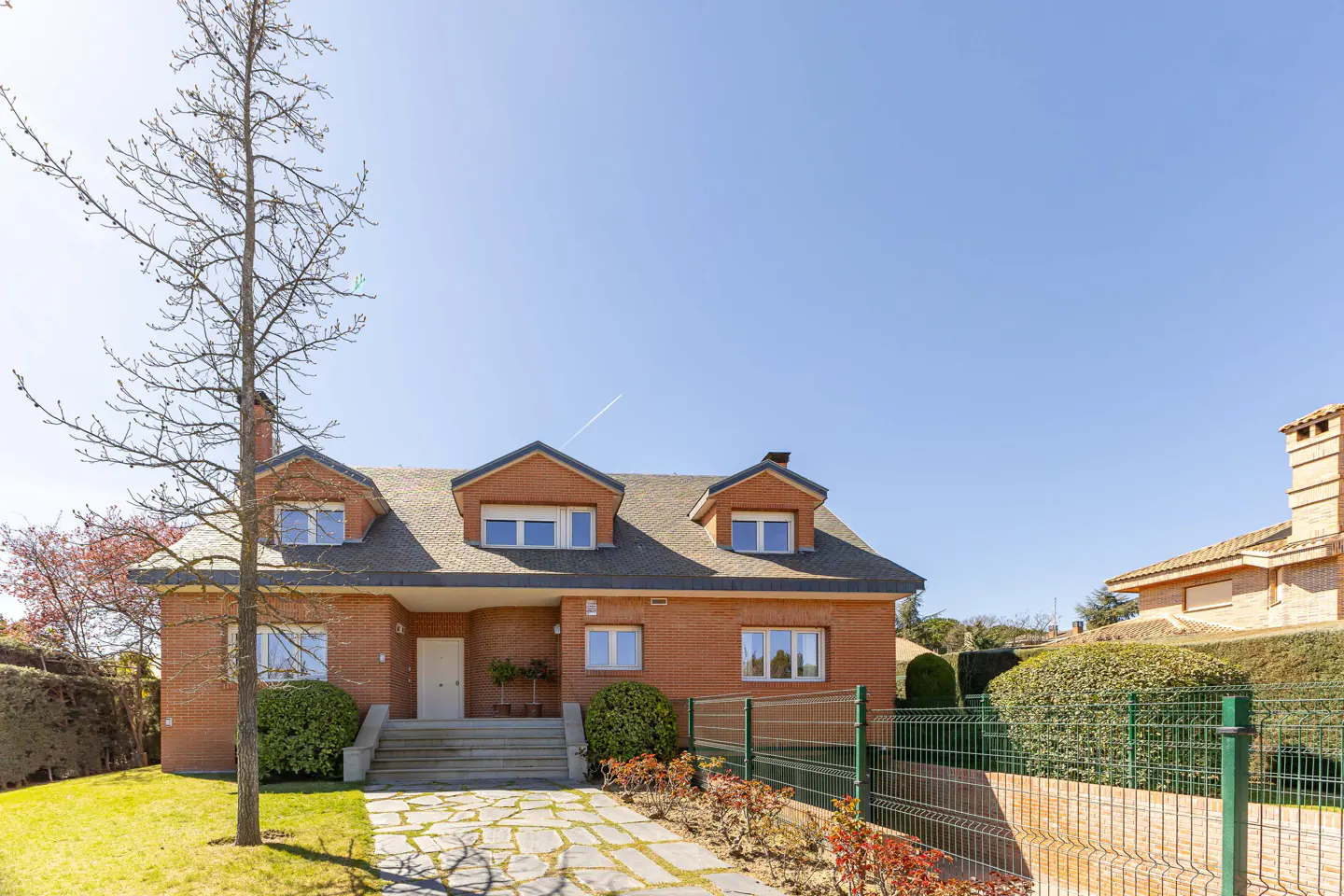 Brick two-story house with dormers, gray roof, and white trim. Stone walkway leads to the front door. A tall tree stands to the left.