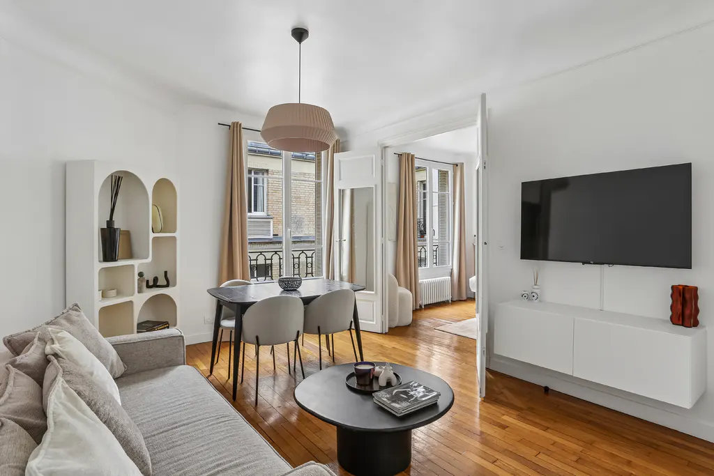 Bright living room with hardwood floors, a grey sofa, a black dining table, a wall-mounted TV, and arched shelving.