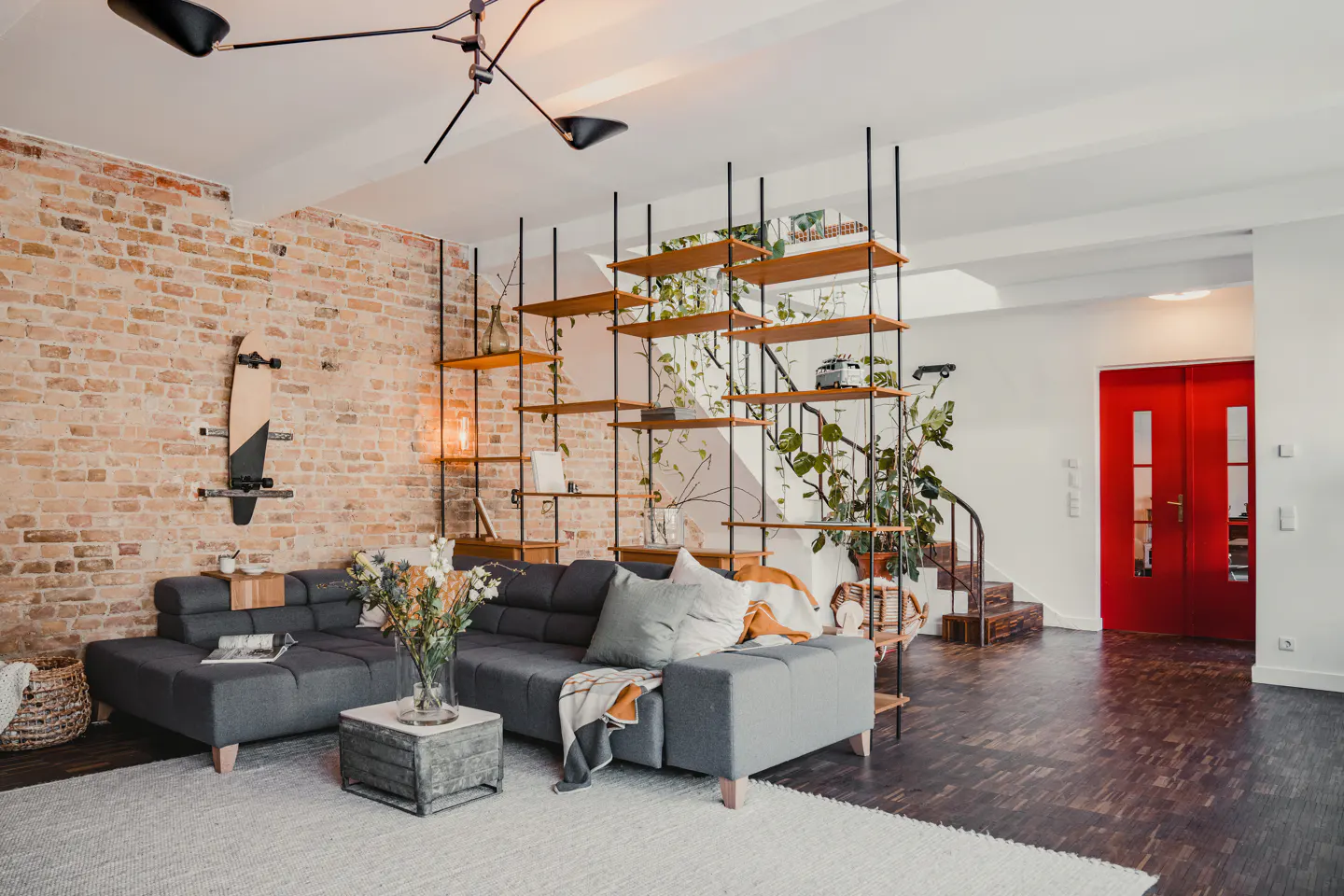 Living room with gray sectional sofa, brick wall with skateboard, and wood shelving unit with plants, leading to a red door.