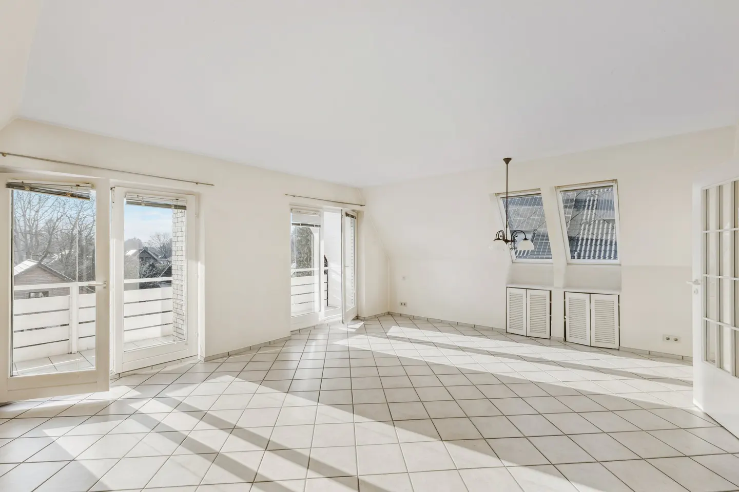 Bright, empty room with white walls, tiled floor, and sunlight streaming through windows and glass doors to balconies.