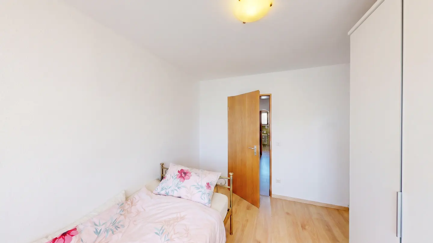 A bright bedroom with white walls, a wood floor, and a single bed with floral bedding. An open wood door leads to another room. A white wardrobe stands against the wall.