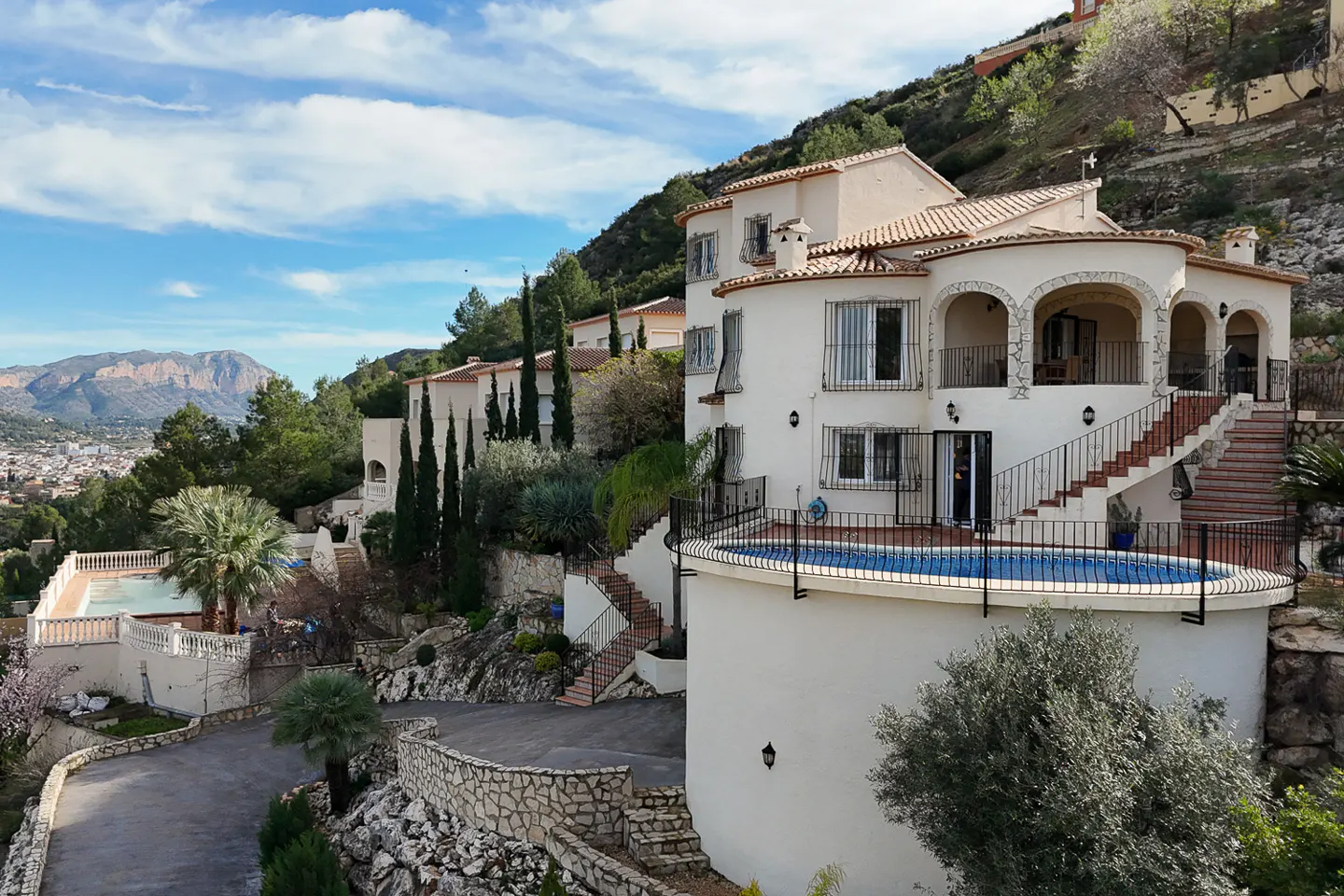 Exterior view of a multi-level white villa with a red tile roof and a pool, set on a hillside with mountain views.