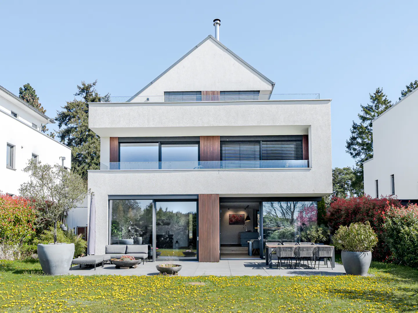Modern white three-story house with a flat roof and a gabled roof, large windows, and a patio with outdoor furniture.