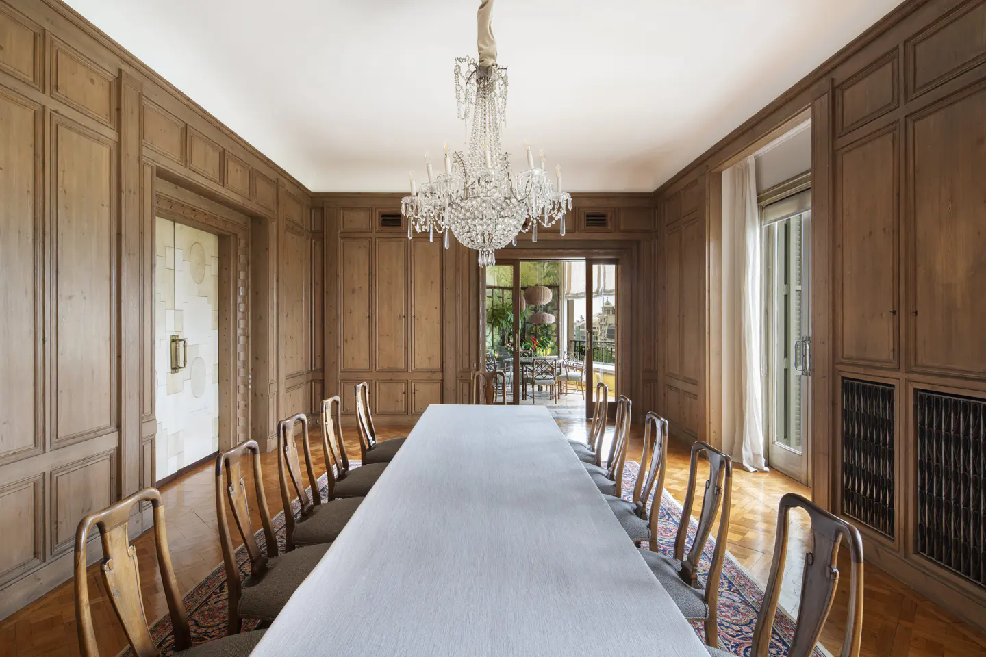 Formal dining room with wood-paneled walls, a long white table, crystal chandelier, and chairs on a patterned rug.