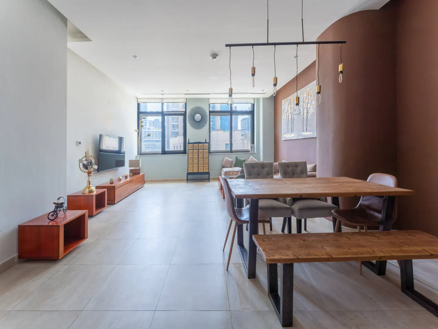 Open-concept living and dining area with a wood table, bench, and chairs under a linear pendant light. Large windows offer city views.