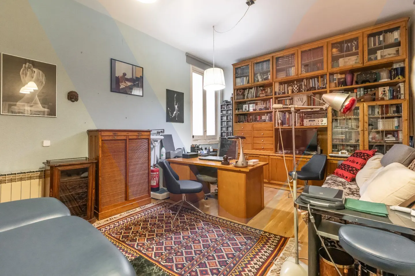 A home office with a large wooden bookcase, desk, chair, and patterned rug. Artwork adorns the light blue walls.