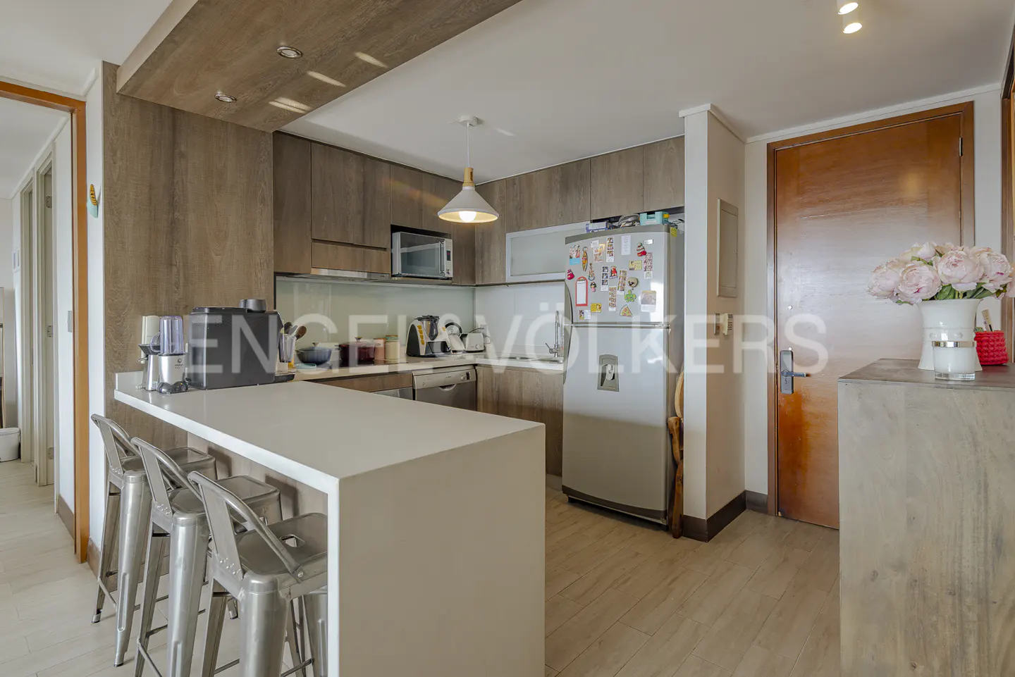 A modern kitchen with wood cabinets, a white island with metal stools, and a stainless steel refrigerator.