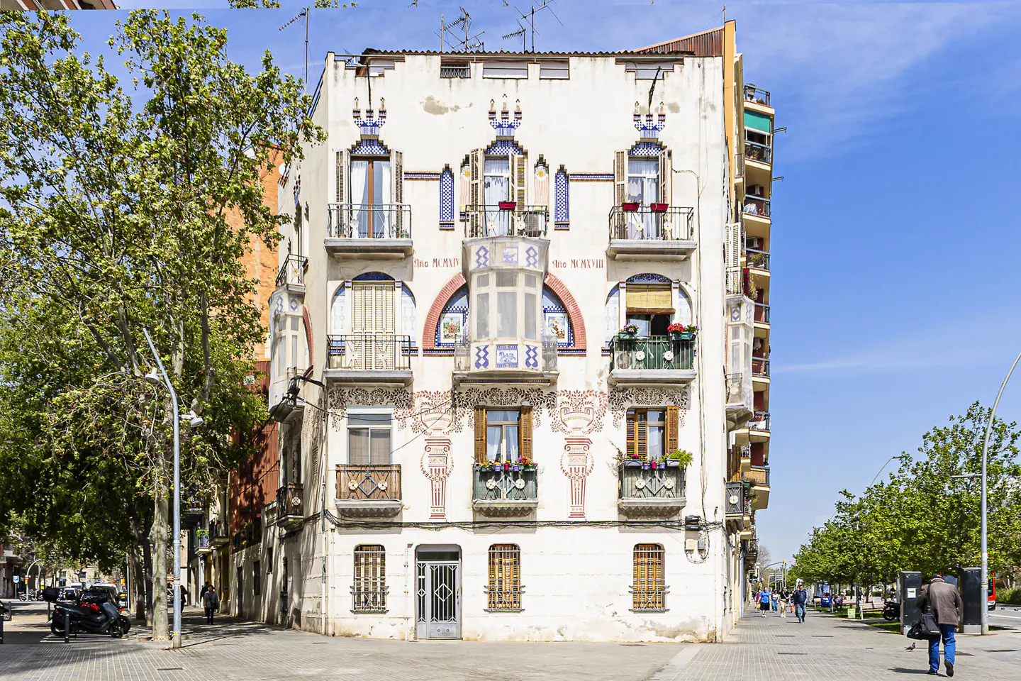 Ornate white building with balconies and decorative tilework, seen from a sunny street with trees and pedestrians.