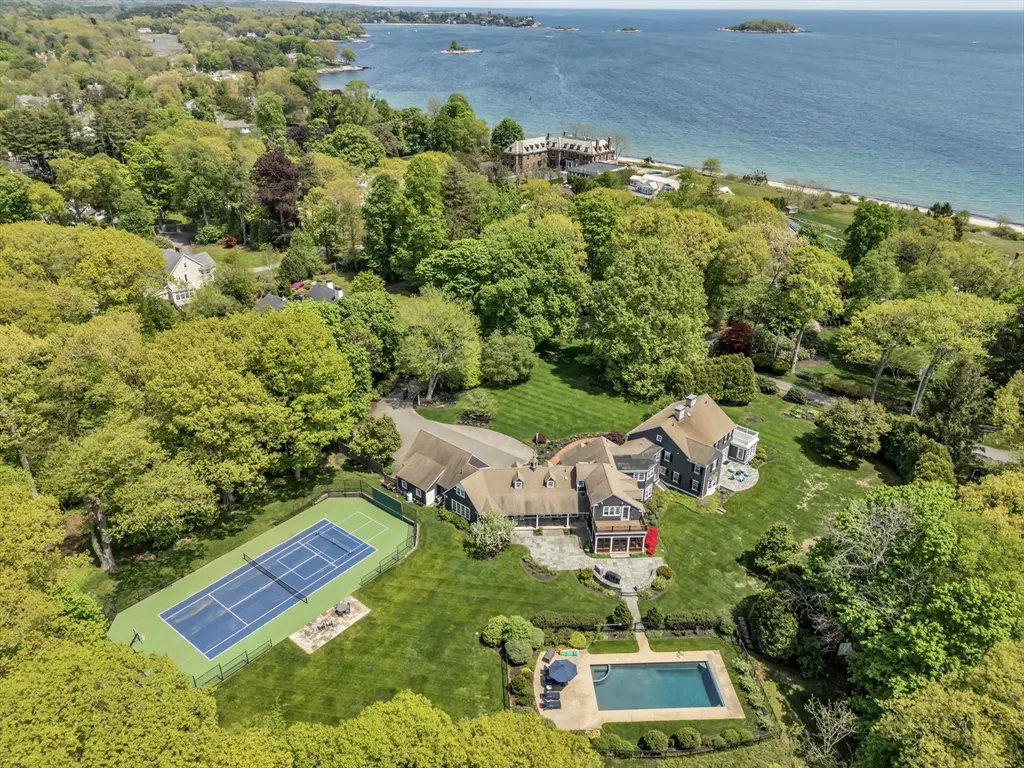 Aerial view of a large gray house with a pool and tennis court, surrounded by green trees and the ocean.