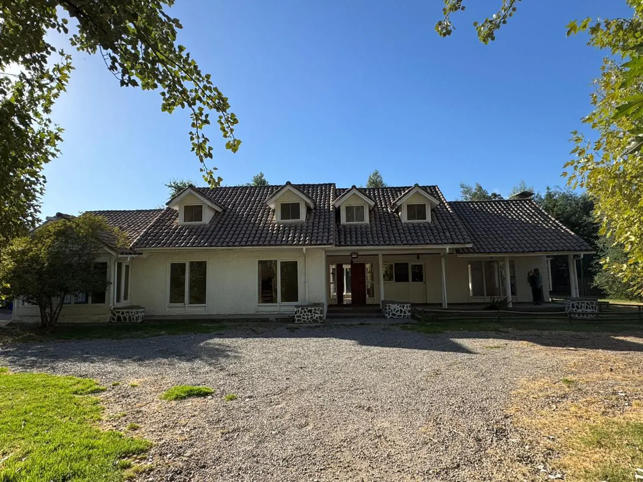Single-story white house with a brown tiled roof, dormer windows, and a large gravel driveway under a clear blue sky.