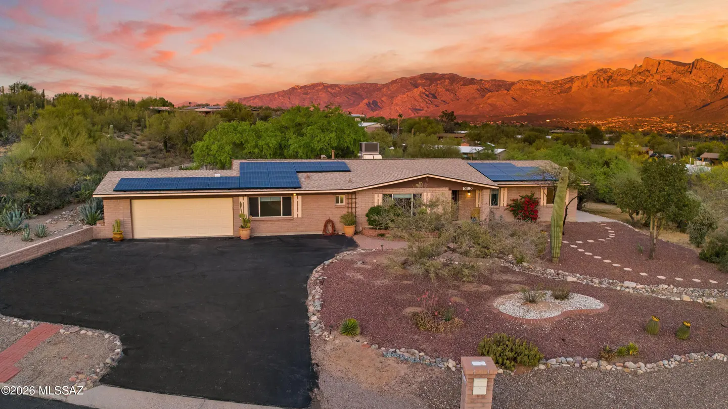 Tan brick home with solar panels, a black driveway, and desert landscaping. Mountains are in the background under a colorful sunset sky.
